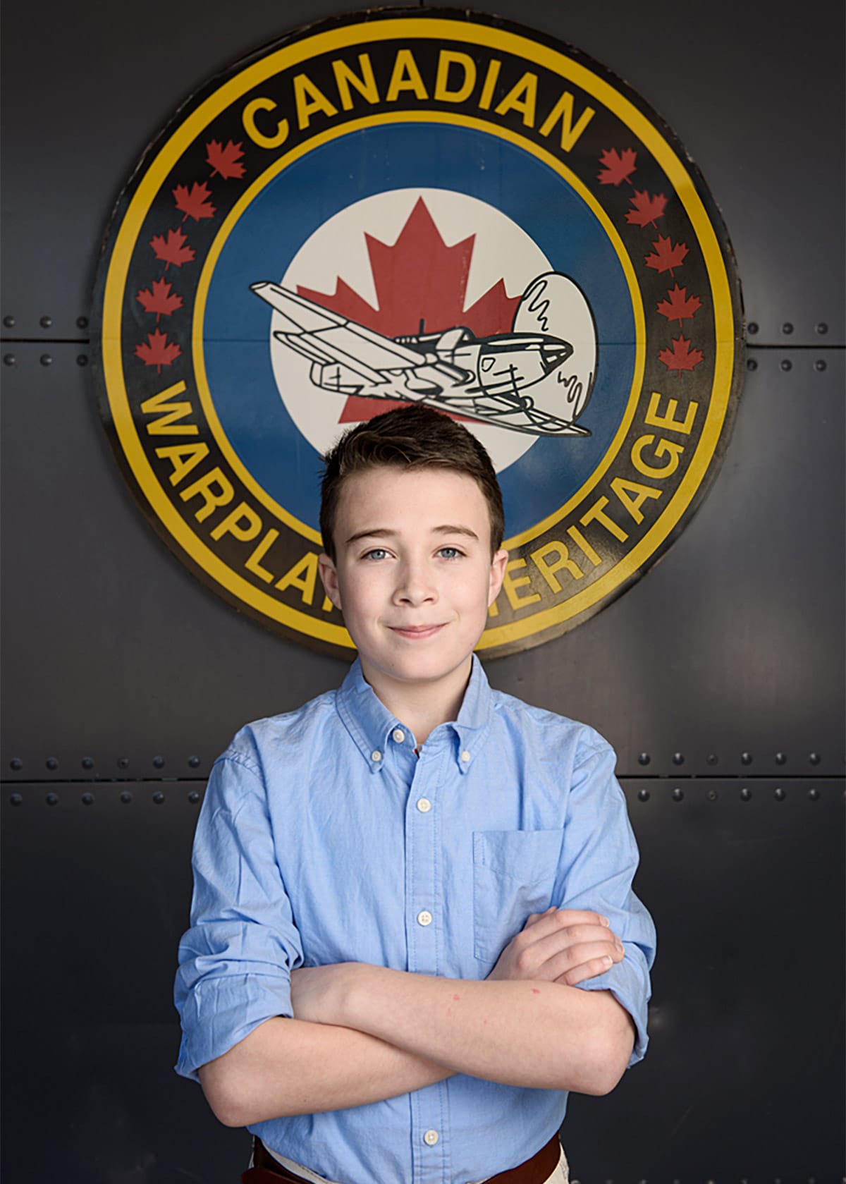 Portrait photograph of a young boy standing cross armed in front of Canadian warplane heritage museum logo